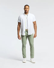 A man stands against a plain background wearing a crisp white Psycho Bunny Gabriel short sleeve shirt, complemented by light olive green pants and pristine white sneakers.