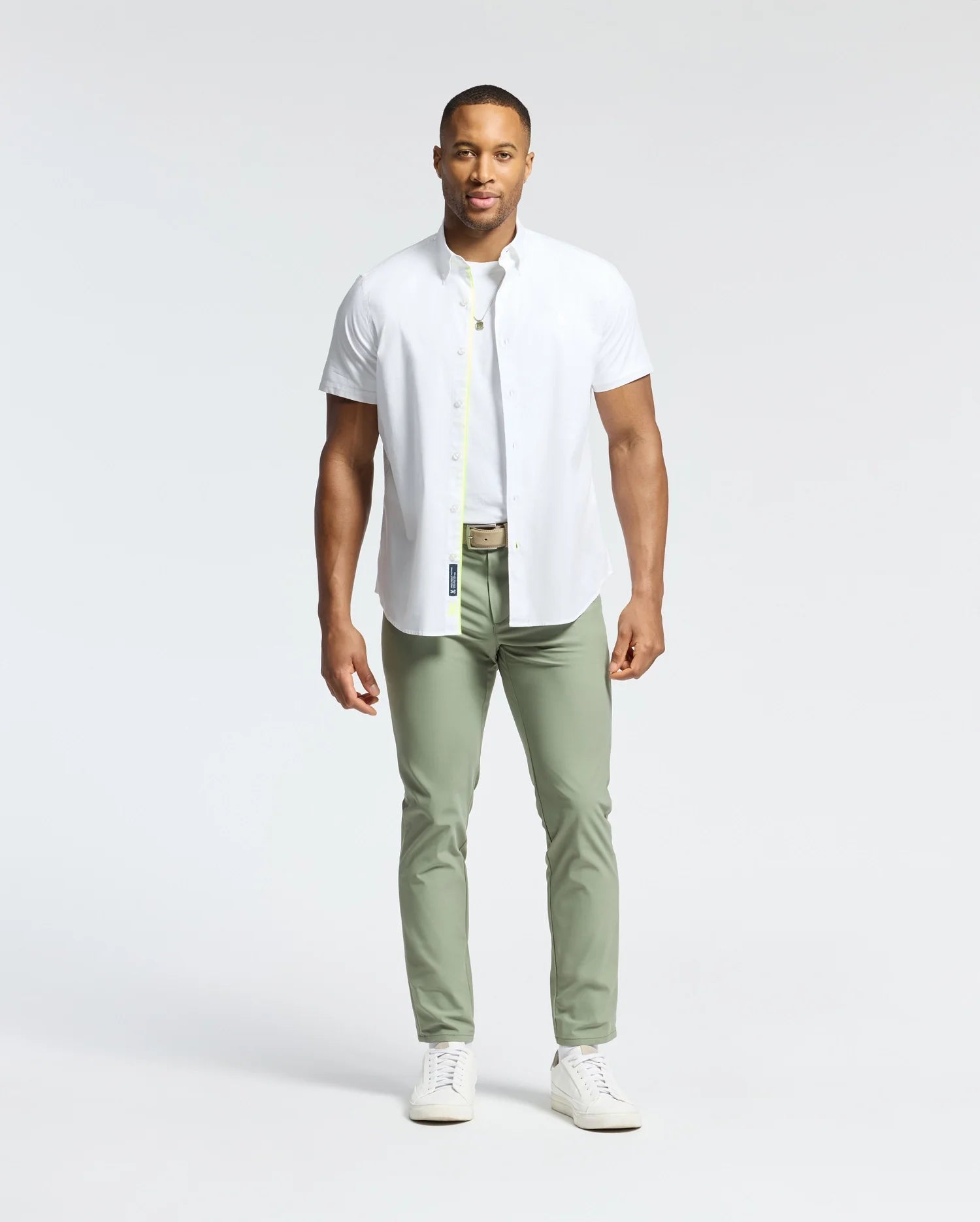A man stands against a plain background wearing a crisp white Psycho Bunny Gabriel short sleeve shirt, complemented by light olive green pants and pristine white sneakers.