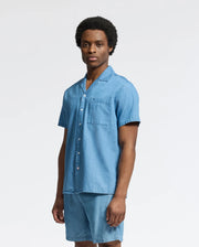 A person wearing the Psycho Bunny MENS KERVIN LIGHTWEIGHT DENIM WOVEN SHIRT (B6Q467F200) with embroidery on the chest is paired with matching shorts. They have short curly hair and stand against a plain white background, slightly turned to the side.
