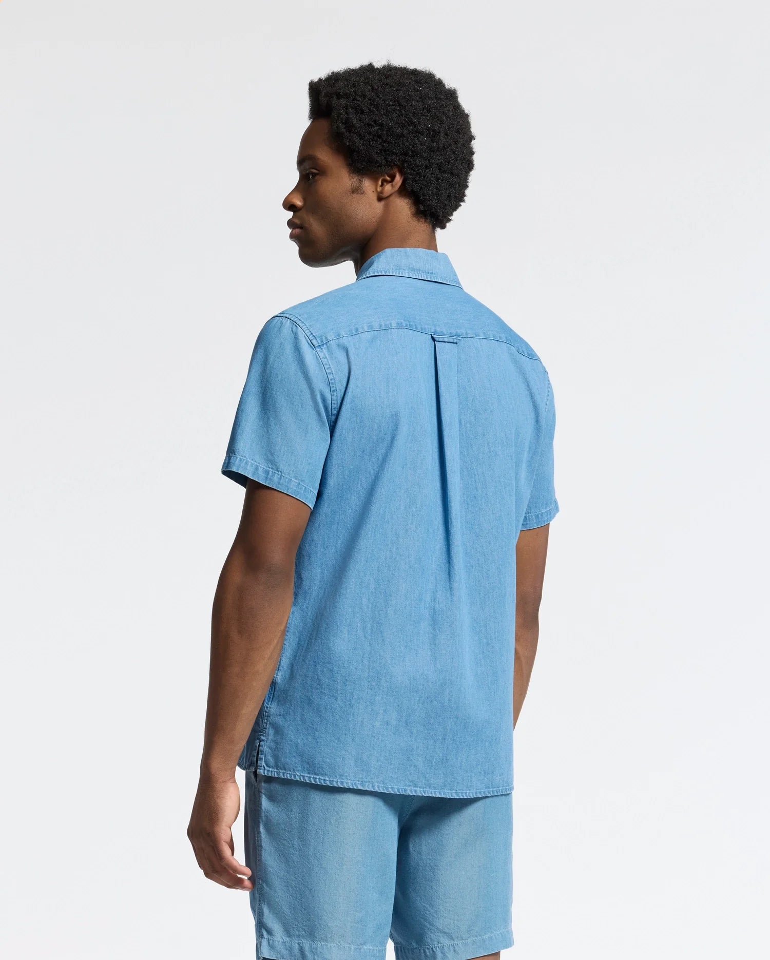 A person with short curly hair stands sideways wearing the Psycho Bunny MENS KERVIN LIGHTWEIGHT DENIM WOVEN SHIRT - B6Q467F200 and matching shorts. This light blue ensemble features subtle chest embroidery, adding elegance against the plain white background.