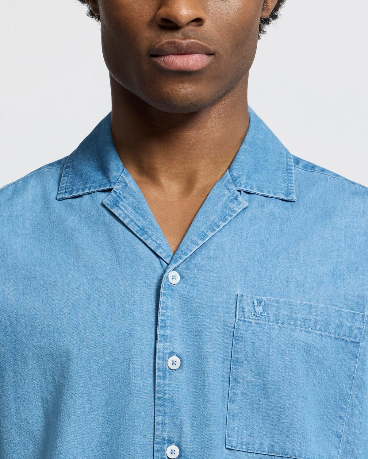 Against a plain background, a person wears the Psycho Bunny MENS KERVIN LIGHTWEIGHT DENIM WOVEN SHIRT (B6Q467F200), showcasing its collar and subtly embroidered chest pocket. Only their torso and part of their face are visible.