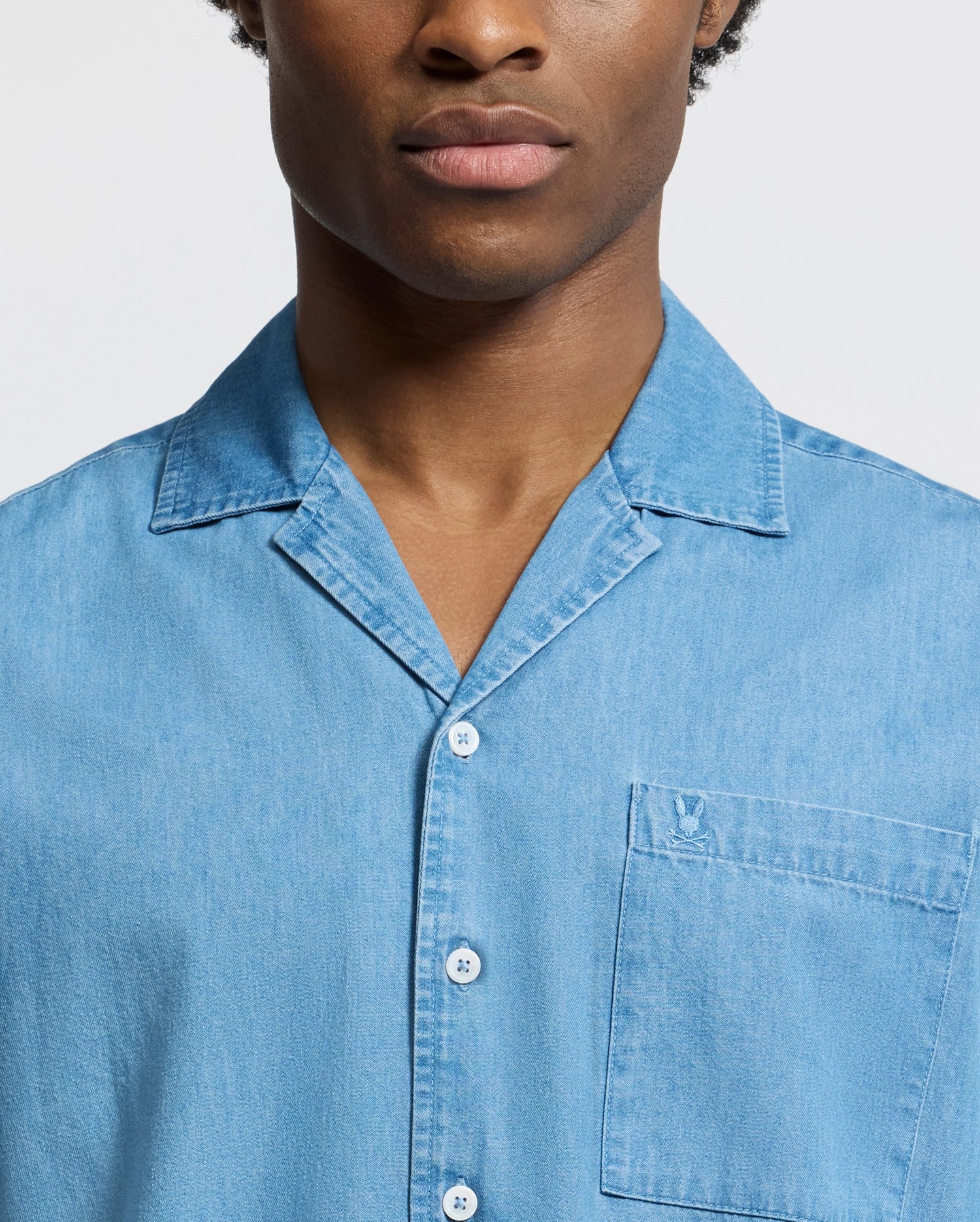 Against a plain background, a person wears the Psycho Bunny MENS KERVIN LIGHTWEIGHT DENIM WOVEN SHIRT (B6Q467F200), showcasing its collar and subtly embroidered chest pocket. Only their torso and part of their face are visible.