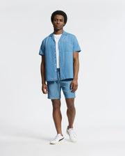 A man stands confidently in a Psycho Bunny MENS KERVIN LIGHTWEIGHT DENIM WOVEN SHIRT over a white T-shirt and denim shorts. Subtle luxury details elevate the casual look, completed with white sneakers against a plain, light background.