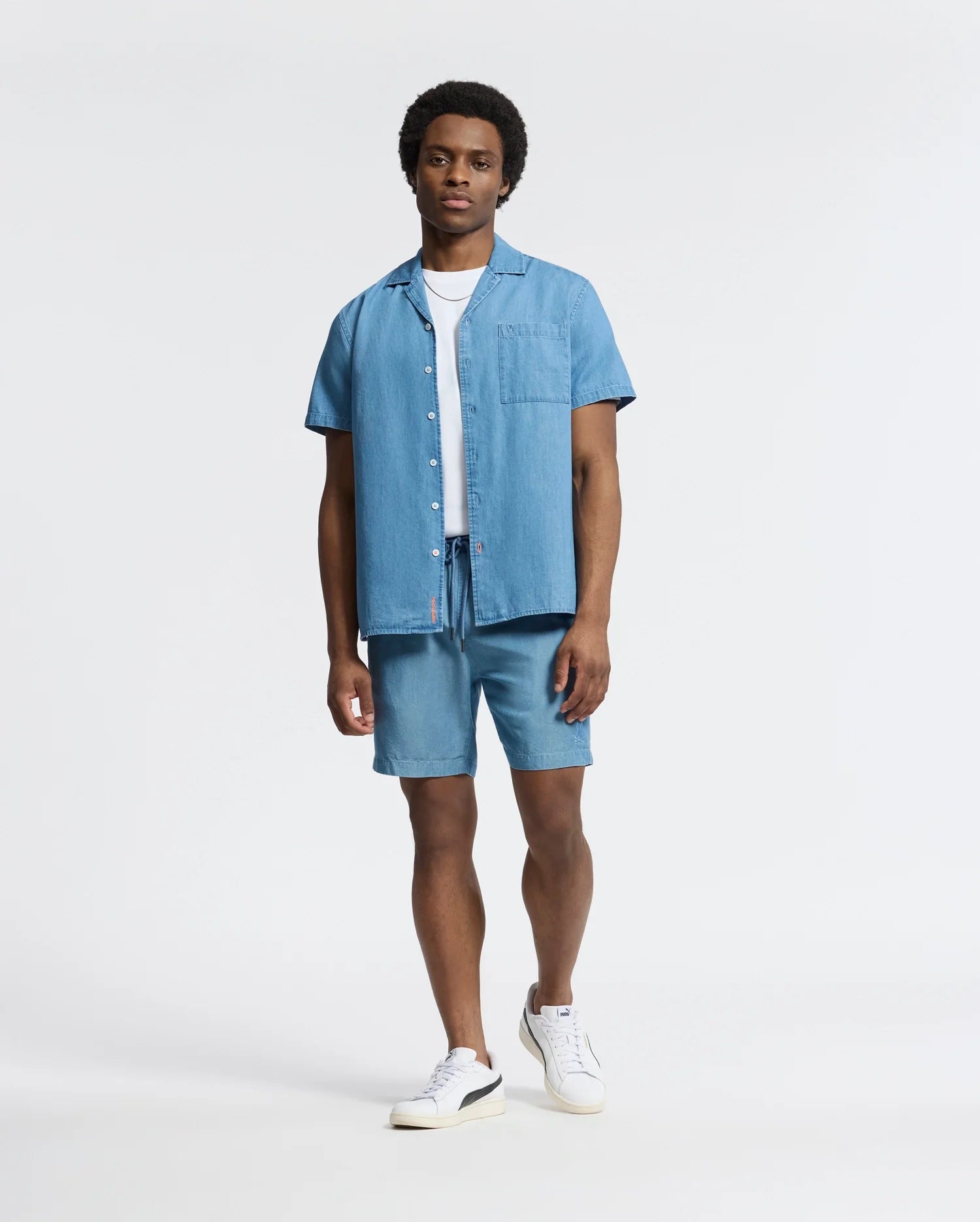 A man stands confidently in a Psycho Bunny MENS KERVIN LIGHTWEIGHT DENIM WOVEN SHIRT over a white T-shirt and denim shorts. Subtle luxury details elevate the casual look, completed with white sneakers against a plain, light background.