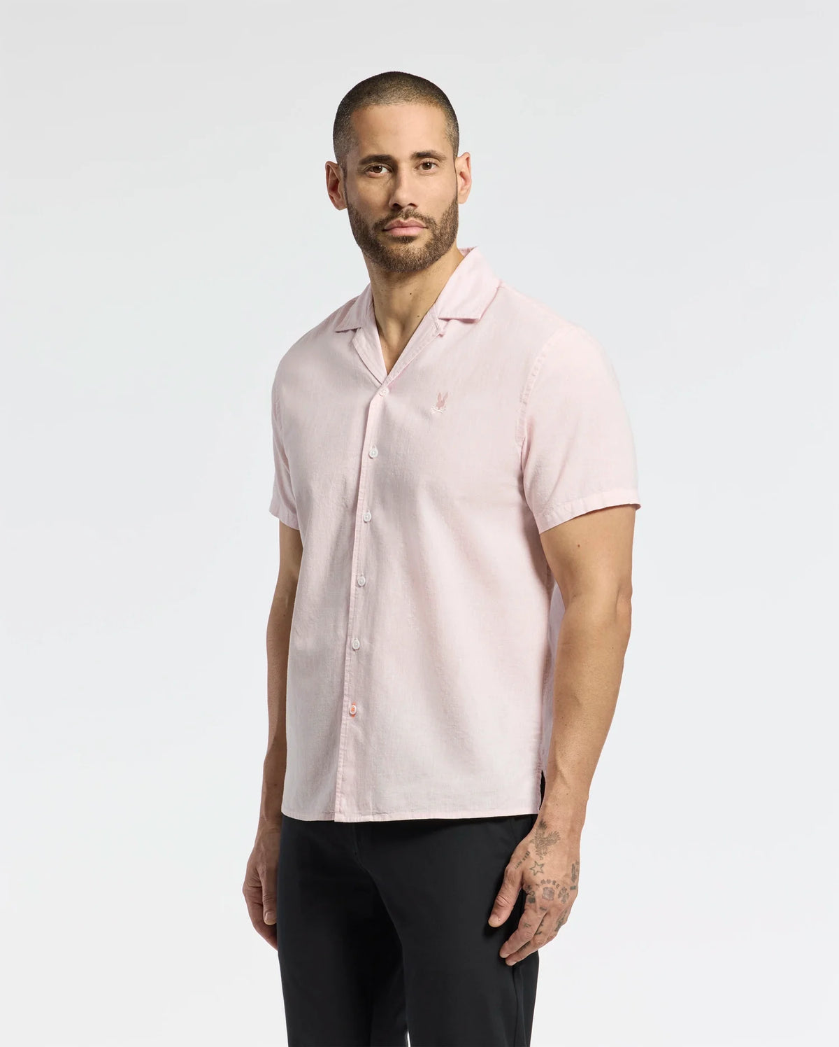A bearded man with short hair wears the Psycho Bunny MENS COBAR WOVEN SHIRT - B6Q522F200, a light pink, striped linen blend short-sleeve button-up, paired with black pants. He stands against a plain white background, gazing at the camera with a neutral expression.