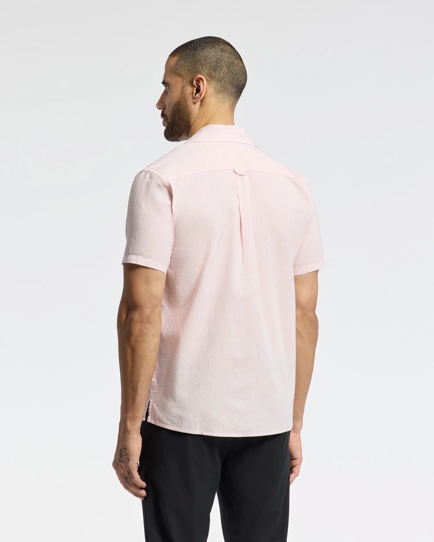 A man with short hair and a beard faces away from the camera, wearing Psycho Bunny's MENS COBAR WOVEN SHIRT (B6Q522F200), a light pink embroidered short-sleeve button-up, paired with black pants against a plain white background.