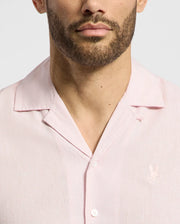 A man with a beard wears the Psycho Bunny MENS COBAR WOVEN SHIRT - B6Q522F200, a light pink linen blend with subtle embroidery on the left side. The cropped shot focuses on his neck and torso against a plain background, highlighting the fabric's texture.