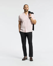 A man confidently stands against a simple backdrop, clad in Psycho Bunny's MENS COBAR WOVEN SHIRT - B6Q522F200, a stylish light pink linen blend short-sleeve shirt paired with black trousers. He holds a black jacket over his shoulder and wears leather shoes, exuding relaxed style.