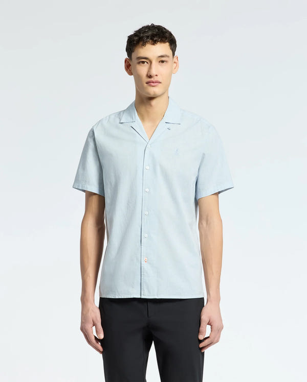A person confidently models a breezy Psycho Bunny light blue MENS COBAR WOVEN SHIRT - B6Q522F200 against a plain white background. With short dark hair, they exude cool composure while gazing into the camera.
