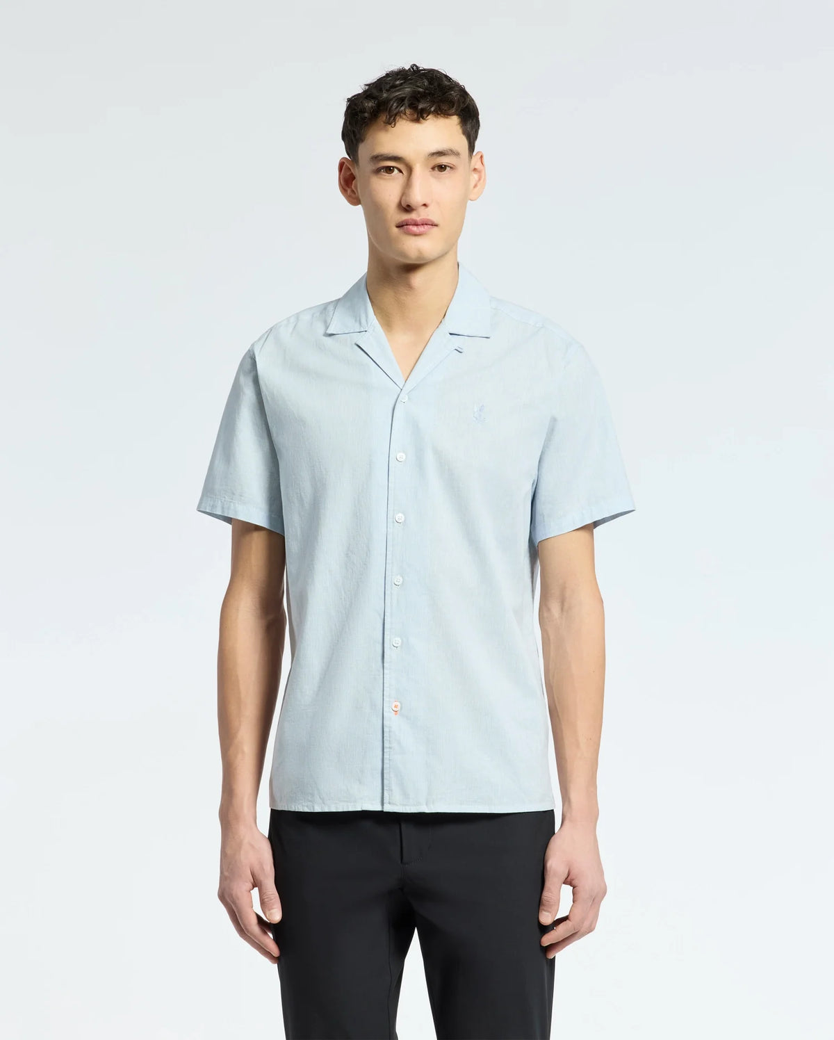 A person confidently models a breezy Psycho Bunny light blue MENS COBAR WOVEN SHIRT - B6Q522F200 against a plain white background. With short dark hair, they exude cool composure while gazing into the camera.