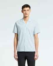 A person confidently models a breezy Psycho Bunny light blue MENS COBAR WOVEN SHIRT - B6Q522F200 against a plain white background. With short dark hair, they exude cool composure while gazing into the camera.
