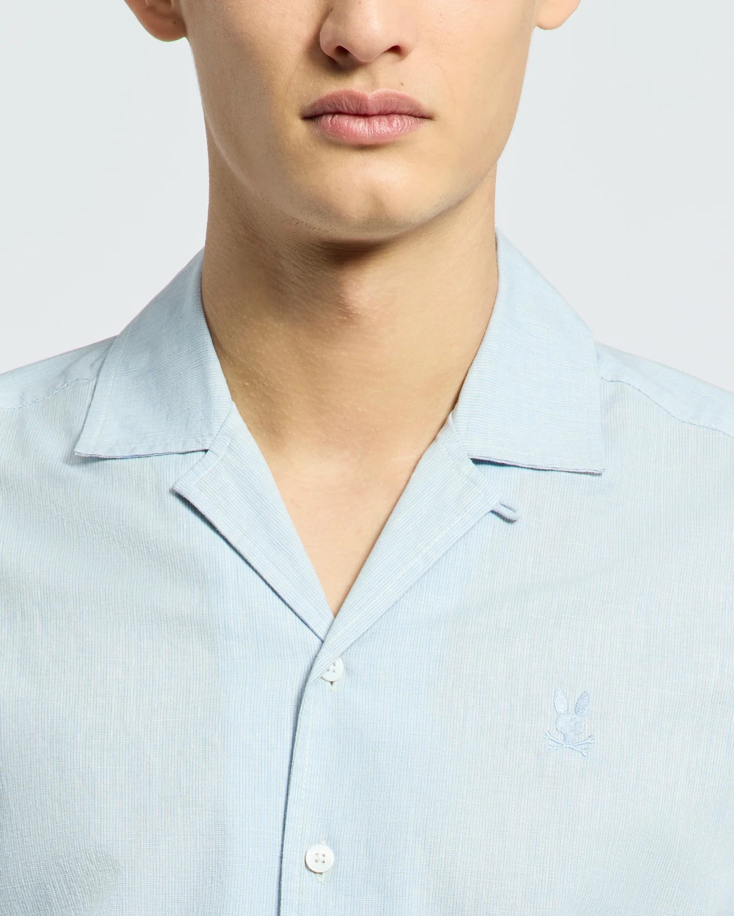 The image shows a person wearing the Psycho Bunny MENS COBAR WOVEN SHIRT - B6Q522F200, a light blue short-sleeve button-up made of cooling fabric featuring a subtle embroidered logo on the chest, with focus on their upper torso and neck against a neutral backdrop.