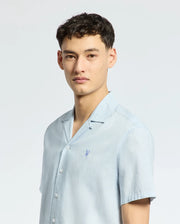 A person with short dark hair is wearing the Psycho Bunny MENS COBAR WOVEN SHIRT - B6Q522F200 in light blue, featuring a small embroidered logo on the chest. The linen blend fabric offers comfort, set against a plain white background.