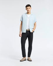 Against a plain background, a person effortlessly captures the beachwear vibe in Psycho Bunny's MENS COBAR WOVEN SHIRT - B6Q522F200, a linen blend light blue short-sleeve button-up over a white T-shirt. Paired with black pants, brown loafers, their short curly hair and watch complete the look.