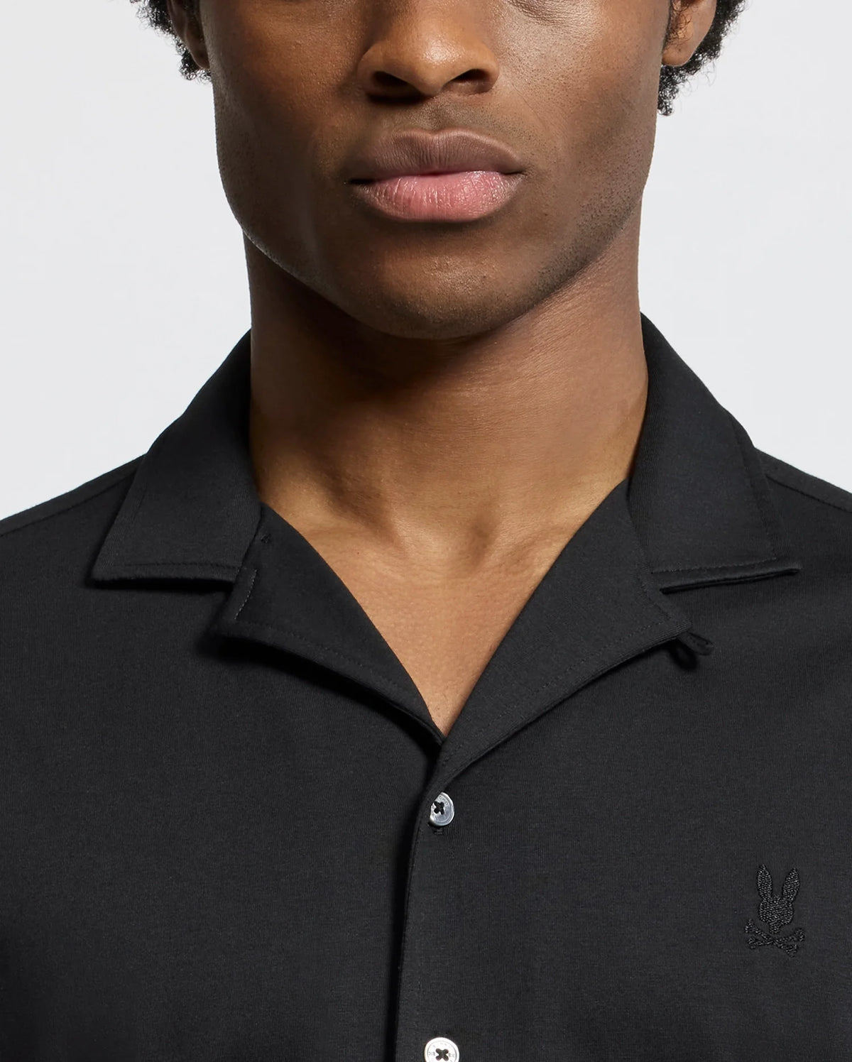 A person is wearing the Psycho Bunny MENS BRYAN CAMP COLLAR SHORT SLEEVE SHIRT (B6Q581F200), which is black and features a small embroidered logo on the chest. The image highlights their upper body against a plain, light-colored background, with their face partially cropped.