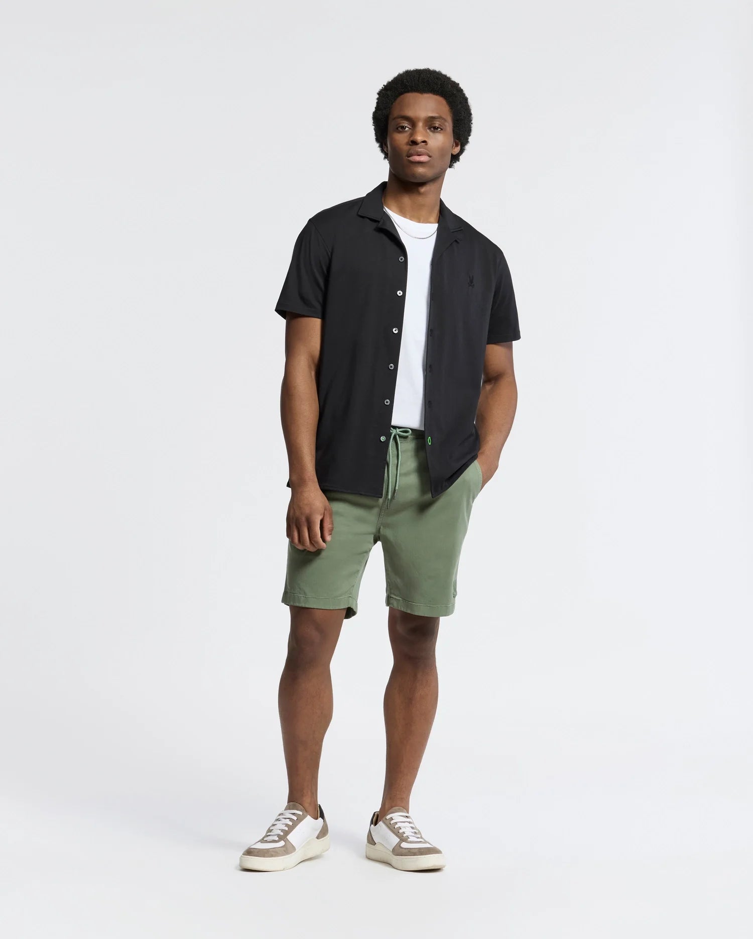 A person stands casually against a plain background, wearing a Psycho Bunny MENS BRYAN CAMP COLLAR SHORT SLEEVE SHIRT - B6Q581F200 in black over a white T-shirt, paired with green shorts and sneakers. One hand is in their pocket as they look slightly to the side.