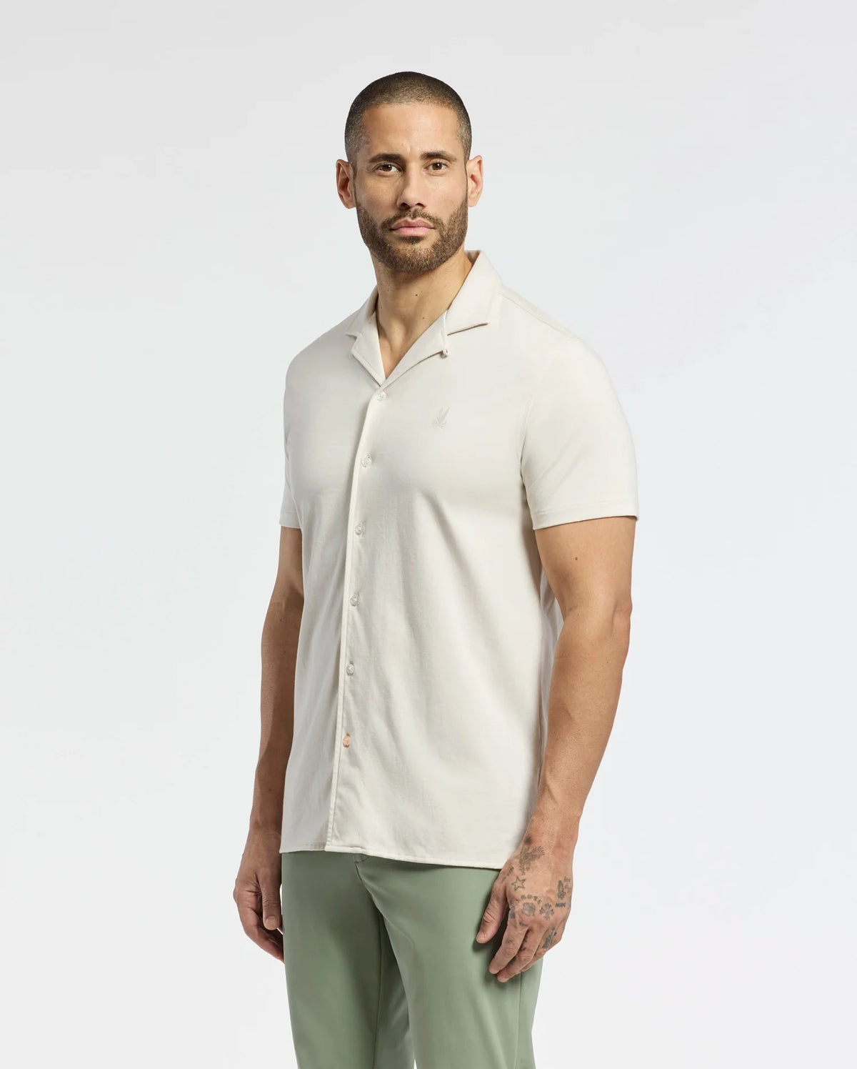 A bearded person with a buzz cut wears the Psycho Bunny MENS BRYAN CAMP COLLAR SHORT SLEEVE SHIRT, crafted from Pima cotton, and light green pants. They stand against a plain white background with one hand by their side.