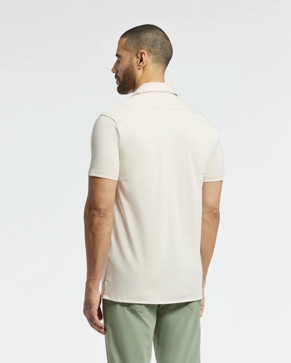 A man with a short beard, facing away, is wearing the light beige Psycho Bunny Men's Bryan Camp Collar Short Sleeve Shirt (B6Q581F200) and green pants against a plain white background.