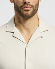 A bearded individual wears the Psycho Bunny MENS BRYAN CAMP COLLAR SHORT SLEEVE SHIRT, B6Q581F200, made from breathable Pima cotton. This light-colored shirt features a subtle chest logo and a stylish camp collar, set against a plain white background.
