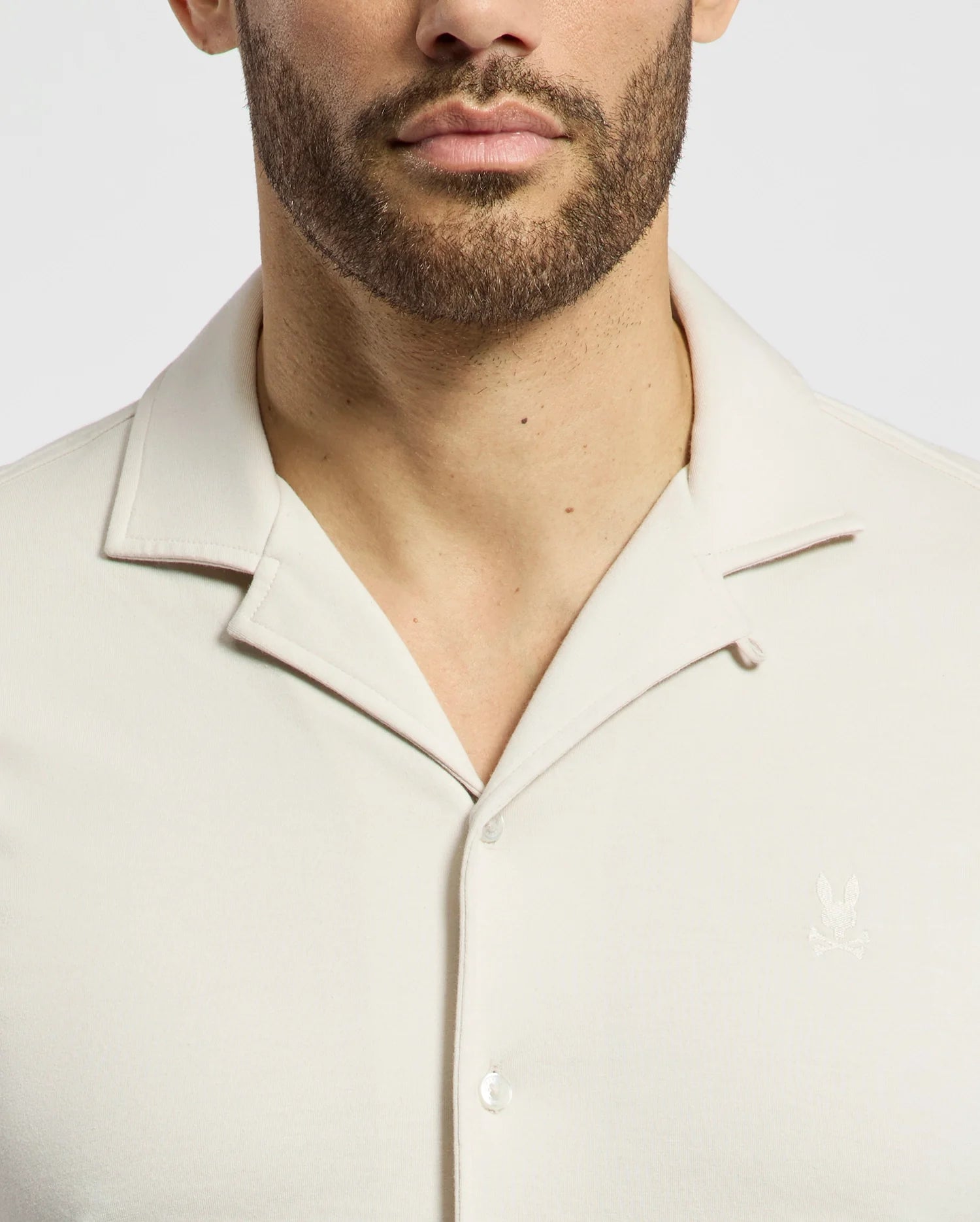 A bearded individual wears the Psycho Bunny MENS BRYAN CAMP COLLAR SHORT SLEEVE SHIRT, B6Q581F200, made from breathable Pima cotton. This light-colored shirt features a subtle chest logo and a stylish camp collar, set against a plain white background.