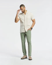 A man wearing Psycho Bunny's beige MENS BRYAN CAMP COLLAR SHORT SLEEVE SHIRT - B6Q581F200, Pima cotton, with green pants adjusts his glasses. He complements the look with brown shoes and a wristwatch, standing against a plain white background and gazing down thoughtfully.