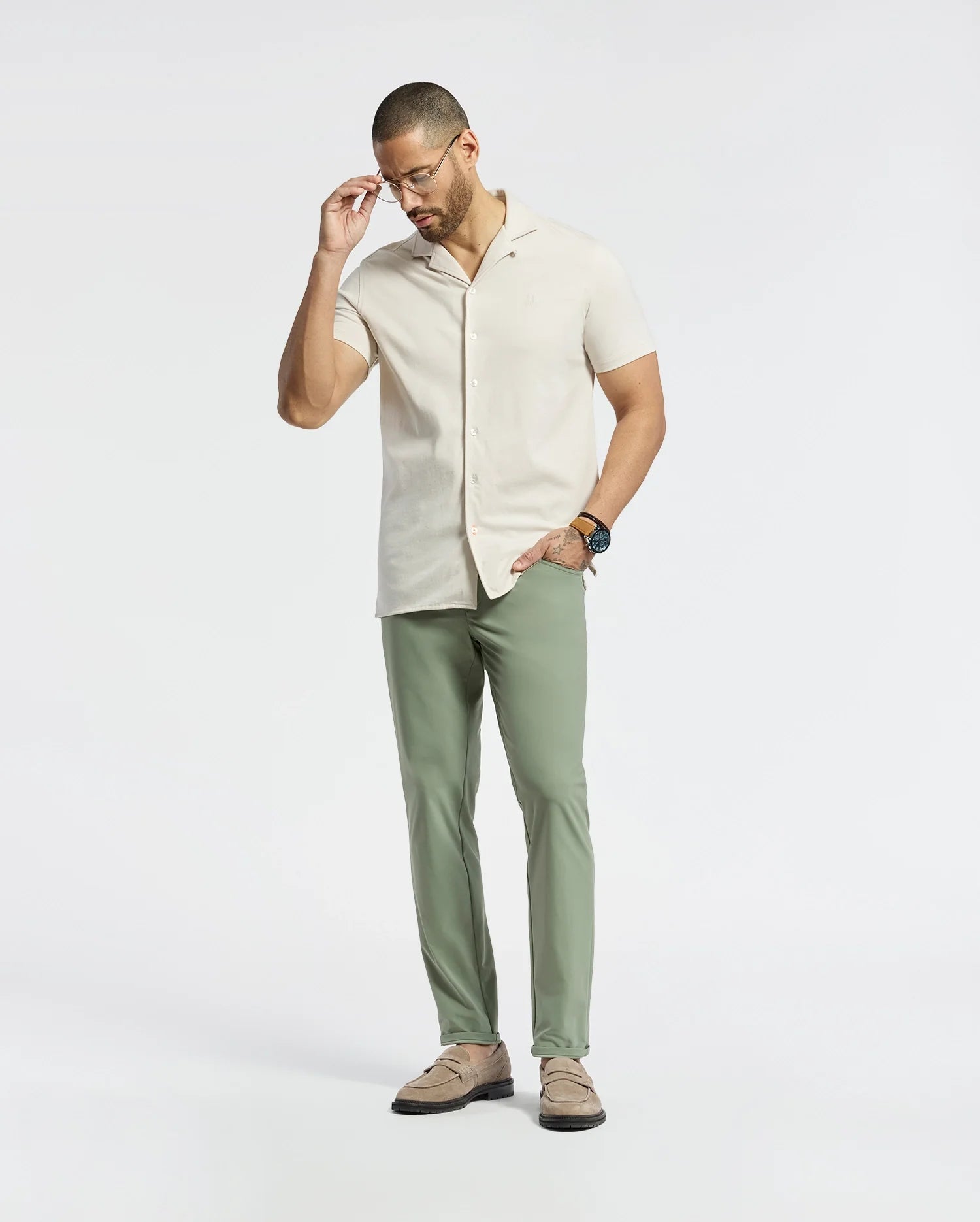 A man wearing Psycho Bunny's beige MENS BRYAN CAMP COLLAR SHORT SLEEVE SHIRT - B6Q581F200, Pima cotton, with green pants adjusts his glasses. He complements the look with brown shoes and a wristwatch, standing against a plain white background and gazing down thoughtfully.