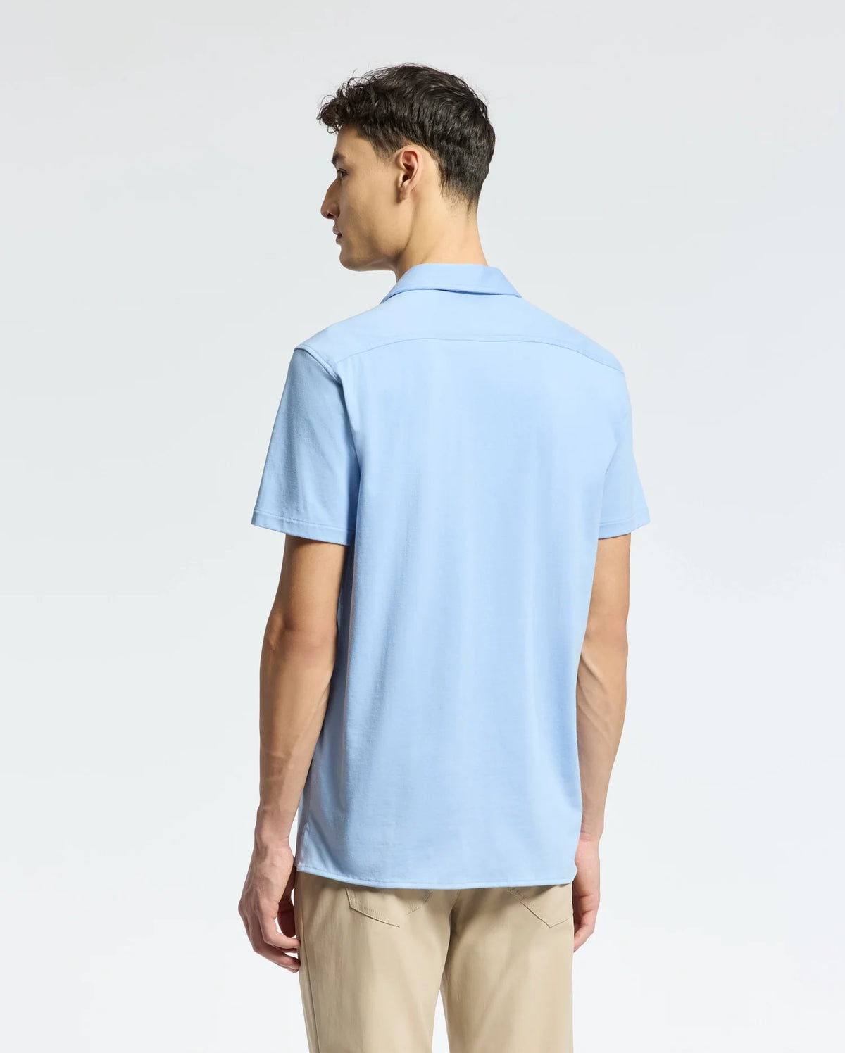 A person with short dark hair faces away, wearing a light blue Men's Bryan Camp Collar Short Sleeve Shirt by Psycho Bunny and beige pants against a plain white background.