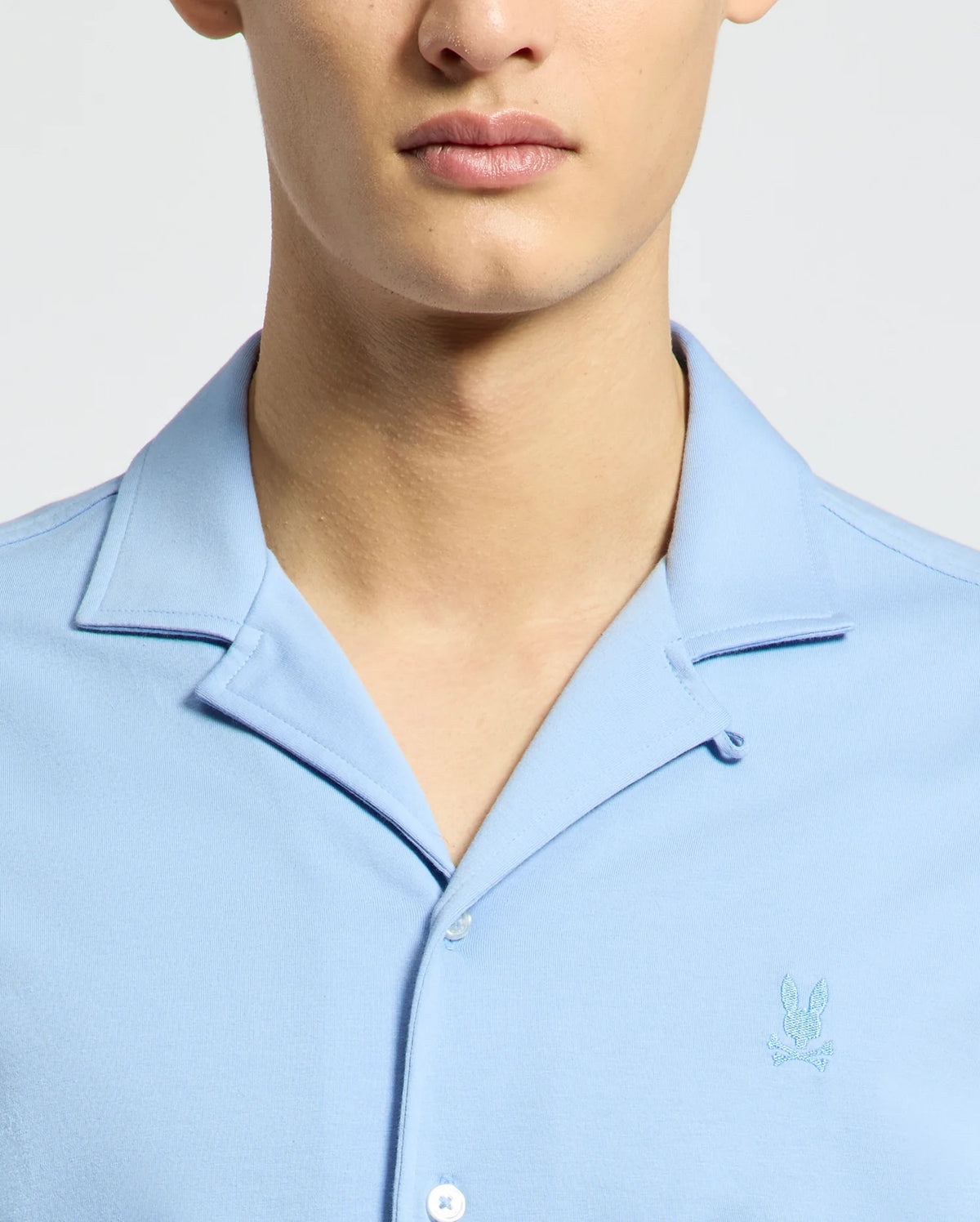 A person wearing the Psycho Bunny MENS BRYAN CAMP COLLAR SHORT SLEEVE SHIRT - B6Q581F200, in light blue and crafted from moisture-wicking performance fabric with an embroidered emblem on the left chest, stands before a neutral background. The focus is on their lower face and torso.