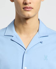 A person wearing the Psycho Bunny MENS BRYAN CAMP COLLAR SHORT SLEEVE SHIRT - B6Q581F200, in light blue and crafted from moisture-wicking performance fabric with an embroidered emblem on the left chest, stands before a neutral background. The focus is on their lower face and torso.