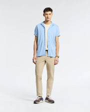 A man stands against a plain backdrop, wearing Psycho Bunny's MENS BRYAN CAMP COLLAR SHORT SLEEVE SHIRT in light blue over a white T-shirt, beige pants, and sneakers. His hands rest by his sides as he gazes slightly left.