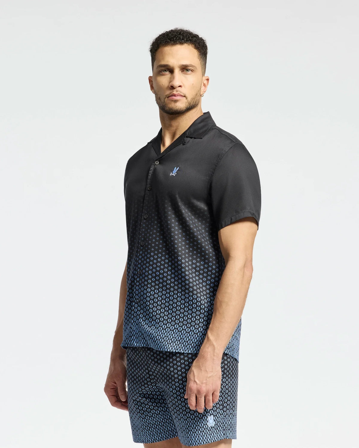 A man stands wearing the Psycho Bunny MENS BENDIGO TENCEL SHIRT in a black and blue ombre design with a camp collar, paired with matching shorts. Made from soft Tencel, his outfit complements his short curly hair and beard as he looks at the camera against a plain white background.