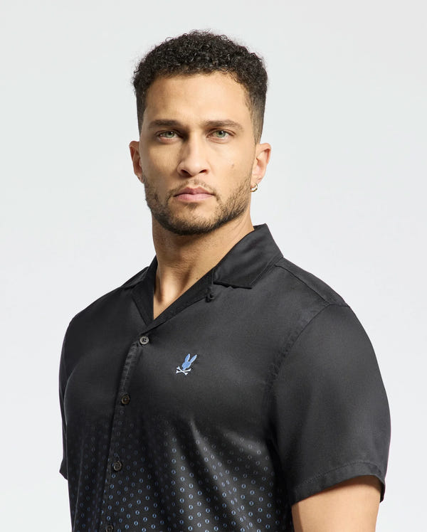 A man with short curly hair and a beard wears the black ombre Psycho Bunny Men's Bendigo Tencel Shirt (B6Q627F200) featuring a camp collar, subtle dotted pattern, and small embroidered emblem. He's turned slightly to his right against a plain background.