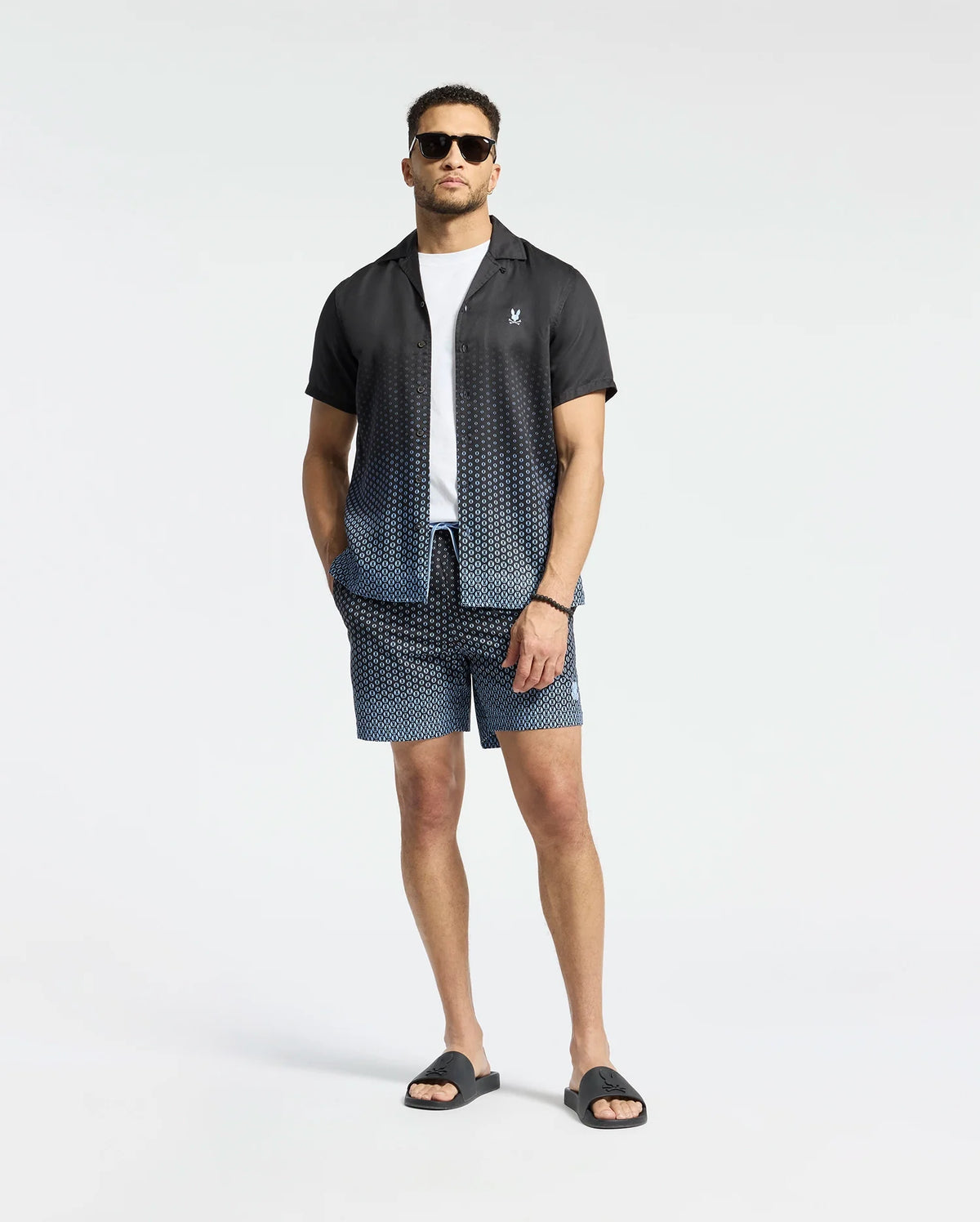 A man wears Psycho Bunny's MENS BENDIGO TENCEL SHIRT, a black and white ombre camp collar shirt over a white T-shirt, paired with matching geometric pattern shorts, black sunglasses, and sandals against a plain white background.