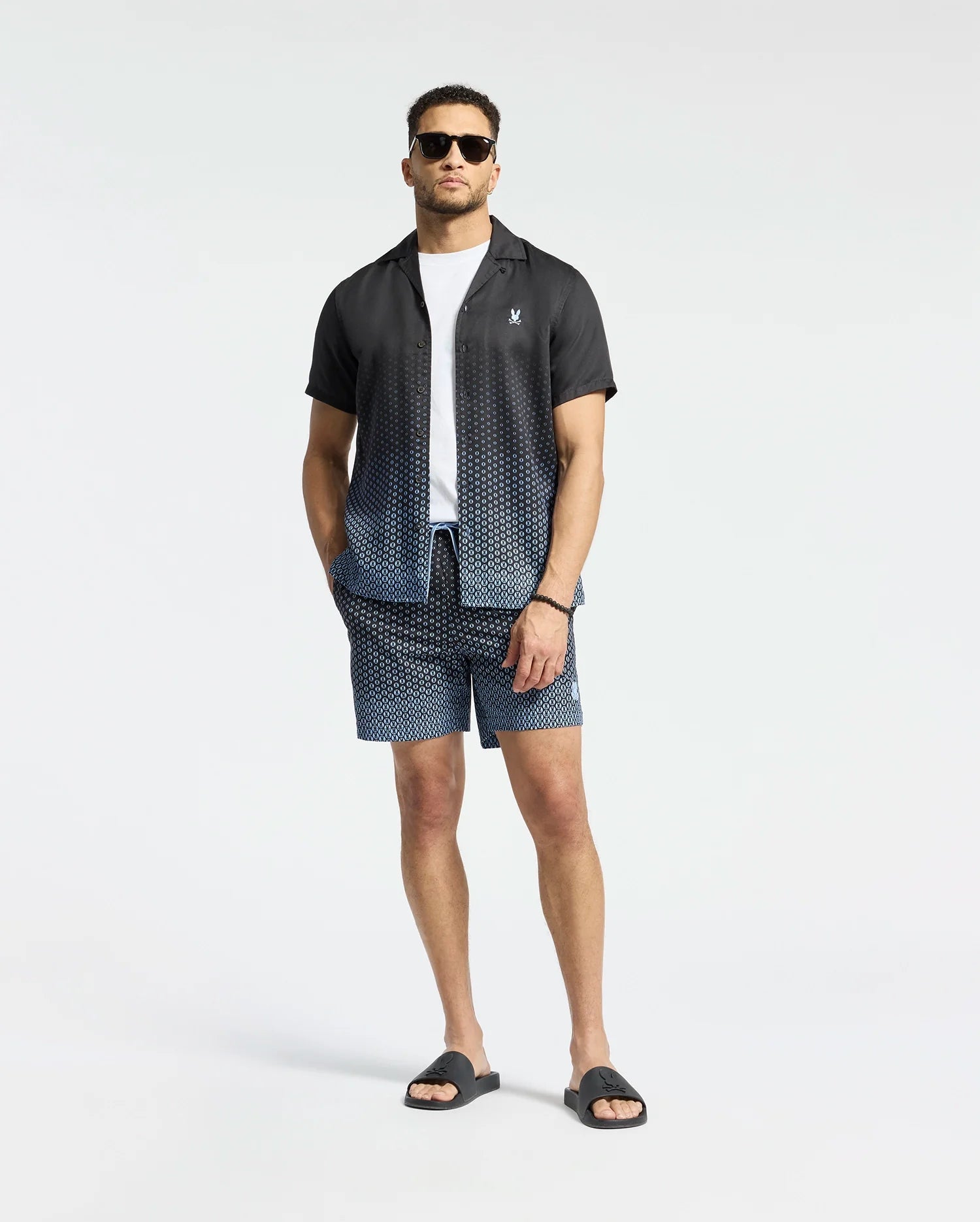 A man wears Psycho Bunny's MENS BENDIGO TENCEL SHIRT, a black and white ombre camp collar shirt over a white T-shirt, paired with matching geometric pattern shorts, black sunglasses, and sandals against a plain white background.