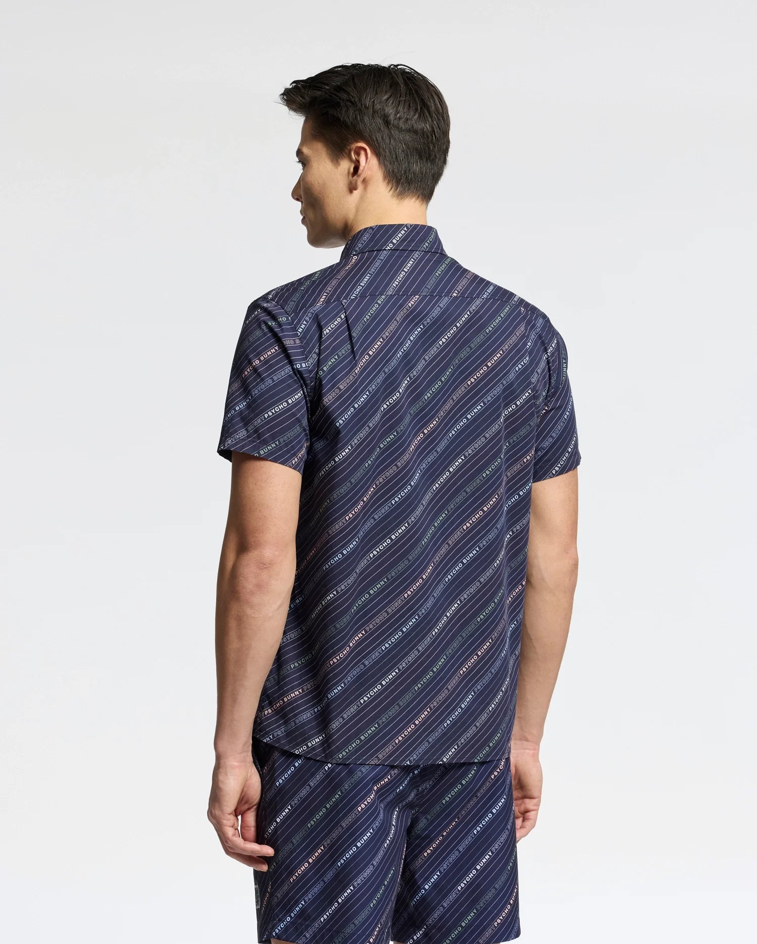 A person stands facing away, wearing a navy MENS RENMARK STRETCH POPLIN WOVEN SHIRT by Psycho Bunny. This premium short-sleeved shirt features diagonal stripes with small white text. Paired with matching shorts and styled with short dark hair, the look is cohesive against a simple background.
