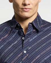 A person is wearing the Psycho Bunny MENS RENMARK STRETCH POPLIN WOVEN SHIRT - B6Q629F200, featuring a dark short sleeve design with multicolored diagonal stripes and "Psycho Bunny" in various colors. The buttoned-up collar stands out against a plain white background.