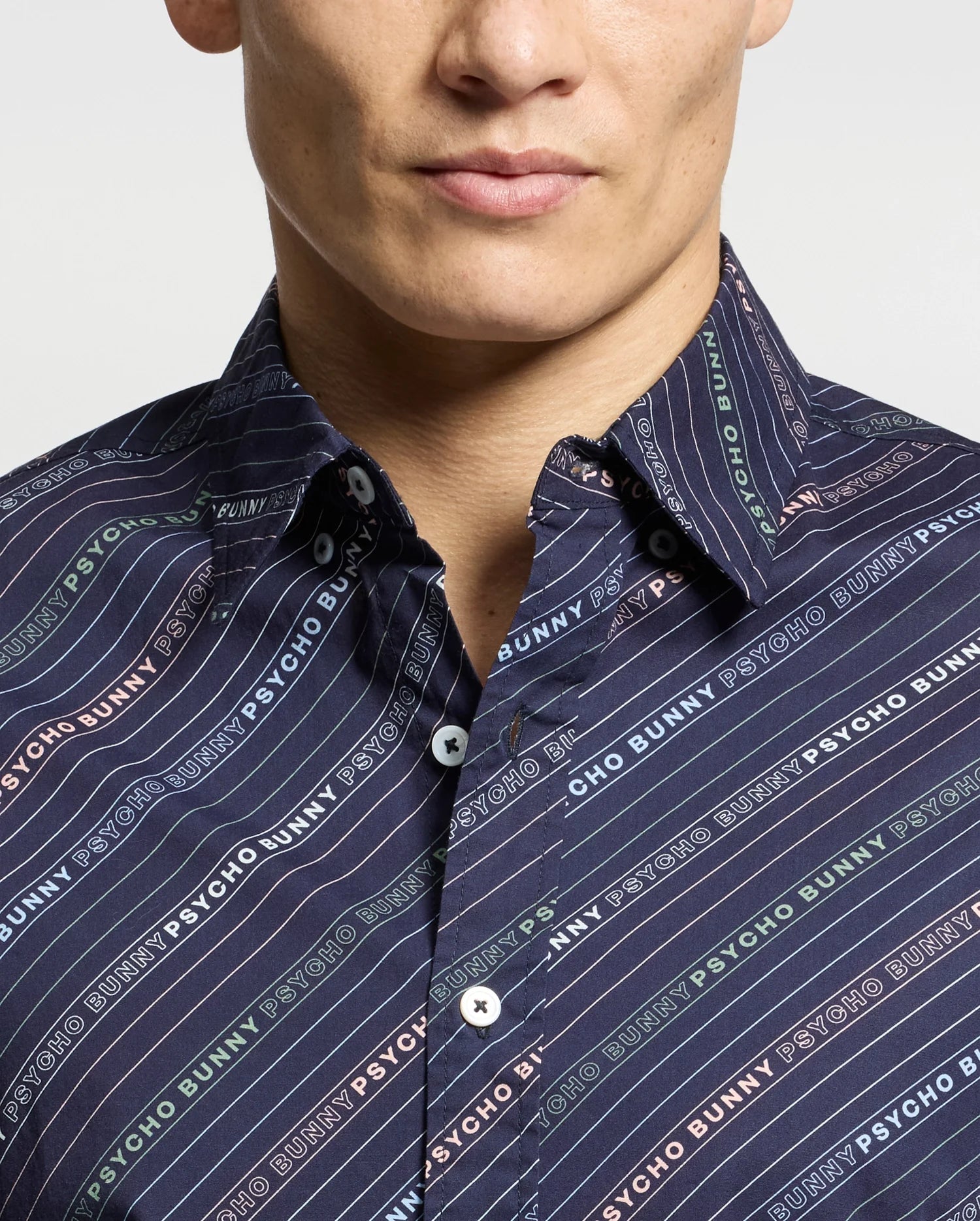 A person is wearing the Psycho Bunny MENS RENMARK STRETCH POPLIN WOVEN SHIRT - B6Q629F200, featuring a dark short sleeve design with multicolored diagonal stripes and "Psycho Bunny" in various colors. The buttoned-up collar stands out against a plain white background.