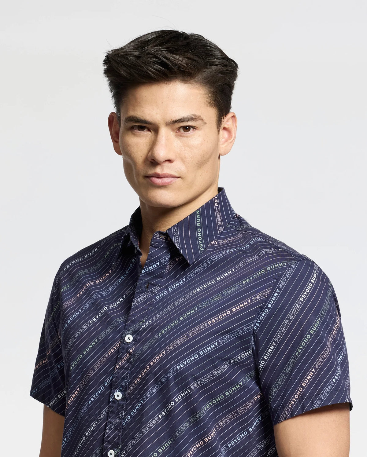 A person with short, dark hair wears a Psycho Bunny MENS RENMARK STRETCH POPLIN WOVEN SHIRT in navy blue. The shirt features multicolored diagonal stripes and text patterns, and has short sleeves against a light background as the person looks toward the camera.