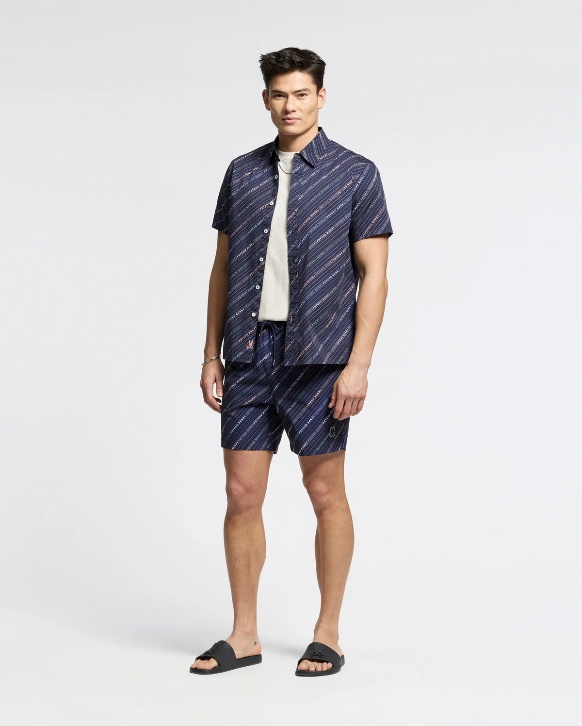 Wearing Psycho Bunny's MENS RENMARK STRETCH POPLIN WOVEN SHIRT - B6Q629F200, a person stands against a plain background. They pair the premium blue short-sleeve button-up with matching subtly striped shorts, layering with a white shirt underneath and finishing the look with dark sandals.