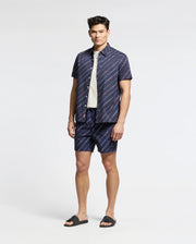 Wearing Psycho Bunny's MENS RENMARK STRETCH POPLIN WOVEN SHIRT - B6Q629F200, a person stands against a plain background. They pair the premium blue short-sleeve button-up with matching subtly striped shorts, layering with a white shirt underneath and finishing the look with dark sandals.