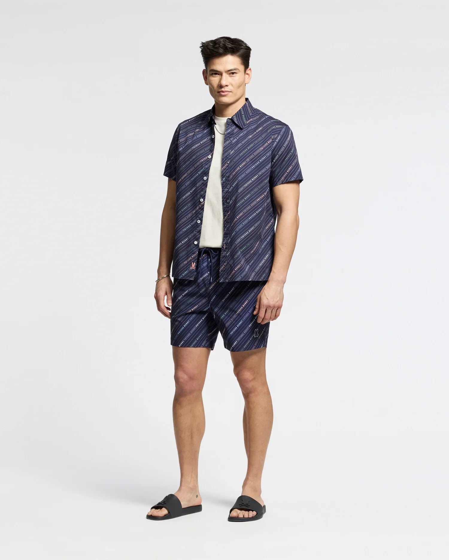 Wearing Psycho Bunny's MENS RENMARK STRETCH POPLIN WOVEN SHIRT - B6Q629F200, a person stands against a plain background. They pair the premium blue short-sleeve button-up with matching subtly striped shorts, layering with a white shirt underneath and finishing the look with dark sandals.