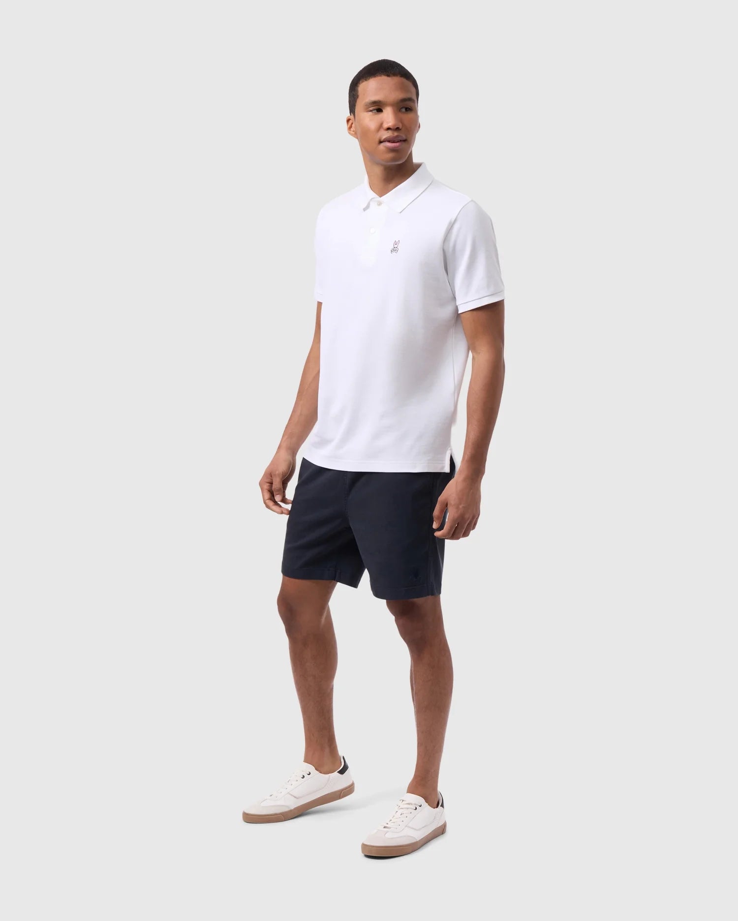 A man wearing a white polo shirt and navy Psycho Bunny eco-friendly shorts standing against a plain background, paired with white sneakers. He looks directly at the camera with a neutral expression.