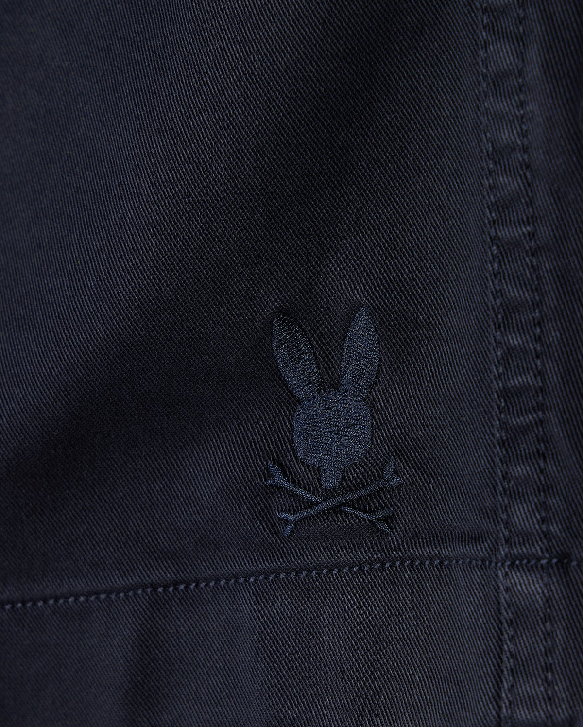 Close-up of a dark fabric texture with a small, embroidered Psycho Bunny logo depicting a stylized rabbit head with crossed bones beneath it on MENS WILLIS STRETCH TENCEL SHORT - B6R239Y1WB.