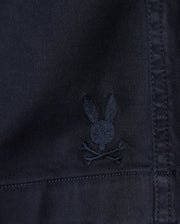 Close-up of a dark fabric texture with a small, embroidered Psycho Bunny logo depicting a stylized rabbit head with crossed bones beneath it on MENS WILLIS STRETCH TENCEL SHORT - B6R239Y1WB.