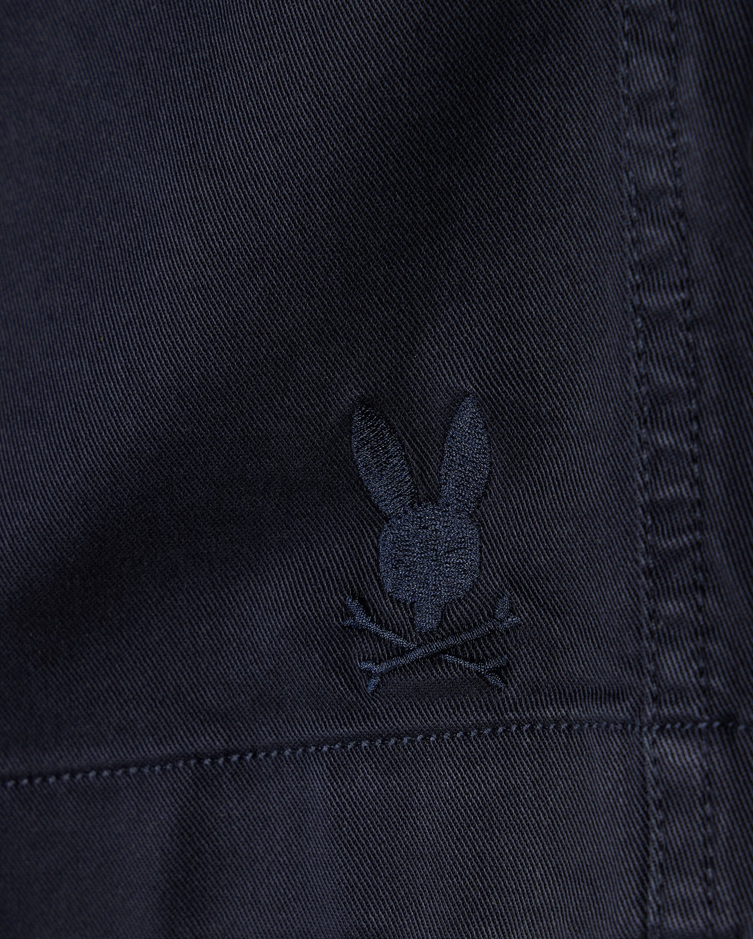 Close-up of a dark fabric texture with a small, embroidered Psycho Bunny logo depicting a stylized rabbit head with crossed bones beneath it on MENS WILLIS STRETCH TENCEL SHORT - B6R239Y1WB.
