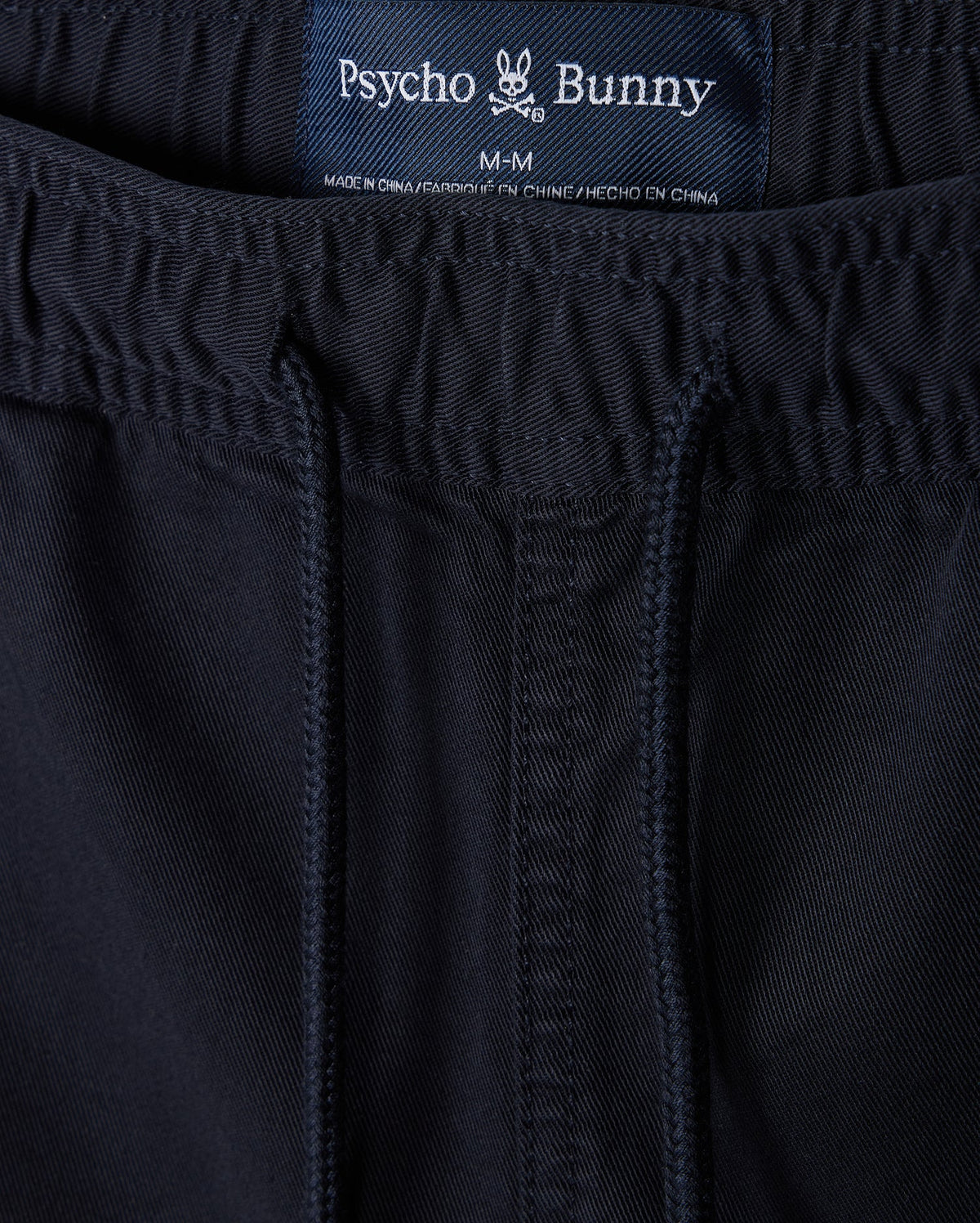 Close-up of navy blue sustainable MENS WILLIS STRETCH TENCEL shorts with a visible label displaying the brand "Psycho Bunny" and size "m-m" sewn into the waistband. The fabric has detailed