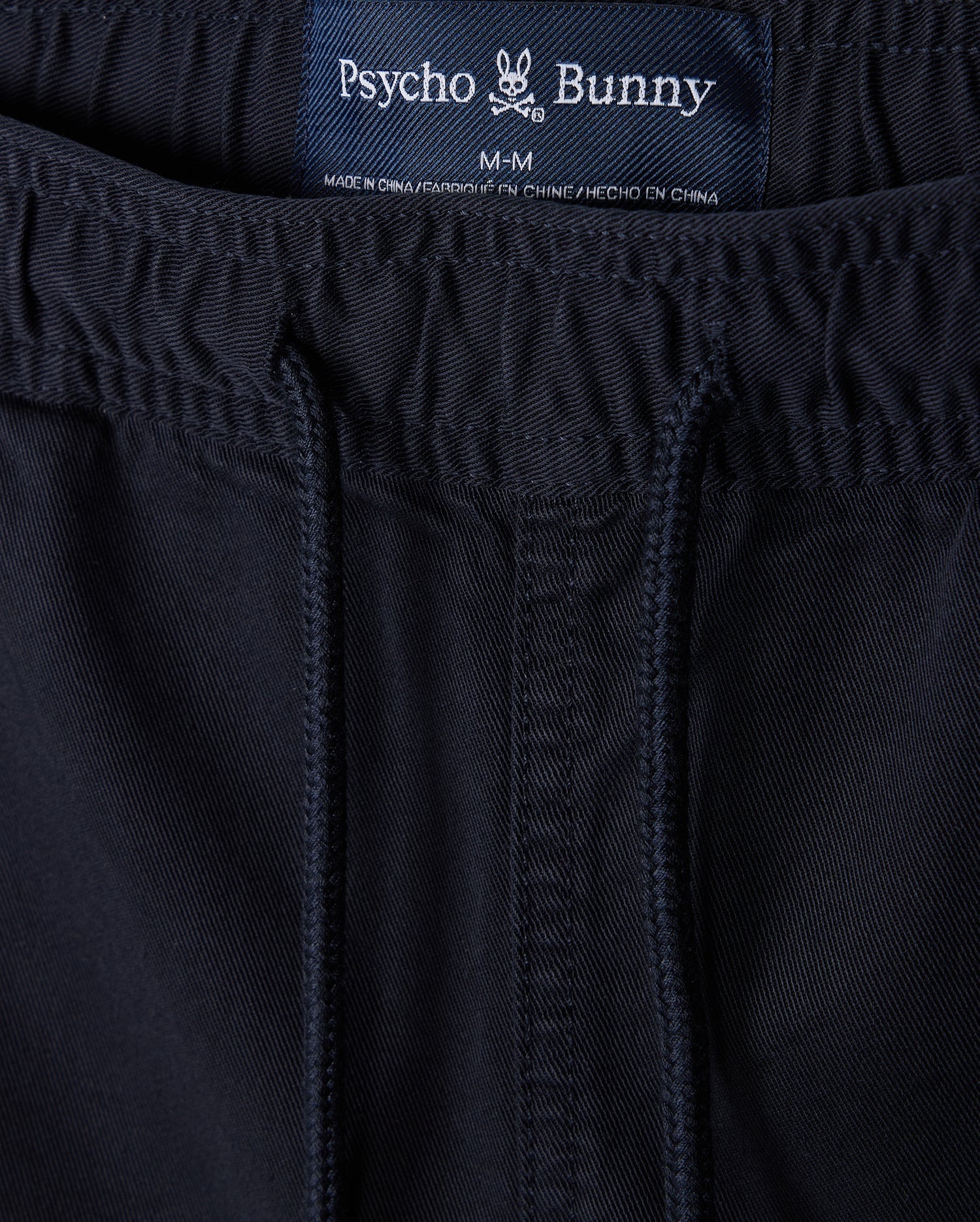 Close-up of navy blue sustainable MENS WILLIS STRETCH TENCEL shorts with a visible label displaying the brand "Psycho Bunny" and size "m-m" sewn into the waistband. The fabric has detailed