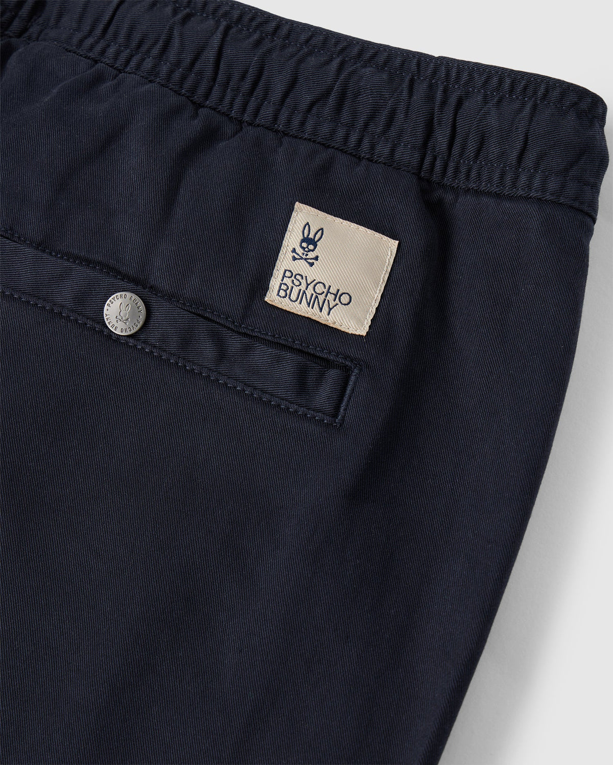 Close-up of Mens Willis Stretch Tencel Shorts from Psycho Bunny, with a detailed view of the waistband, featuring a stylish silver button and a rectangular designer label reading "psycho bunny bunkyo".