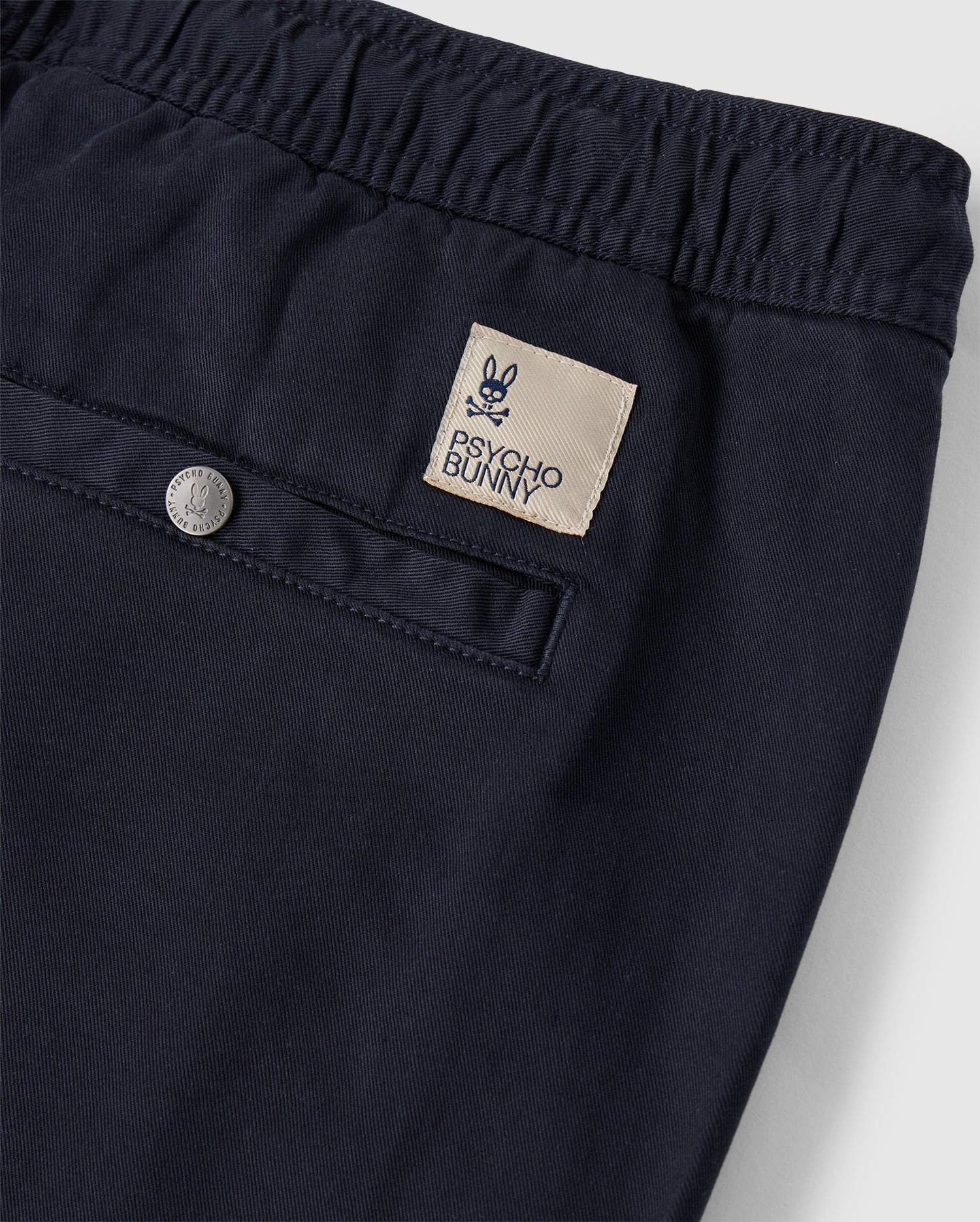Close-up of Mens Willis Stretch Tencel Shorts from Psycho Bunny, with a detailed view of the waistband, featuring a stylish silver button and a rectangular designer label reading "psycho bunny bunkyo".