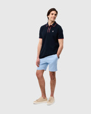 A man stands smiling, dressed in a dark polo shirt with an embroidered Psycho Bunny logo, light blue MENS WILLIS STRETCH TENCEL SHORT - B6R239Y1WB, and cream sneakers, against a plain white background.